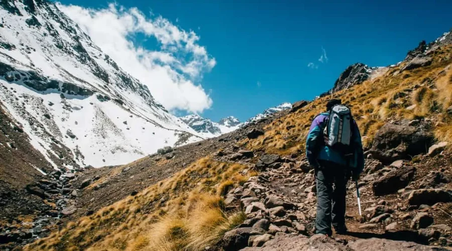 A man with a backpack ascends a mountain trail, surrounded by scenic natural landscapes.