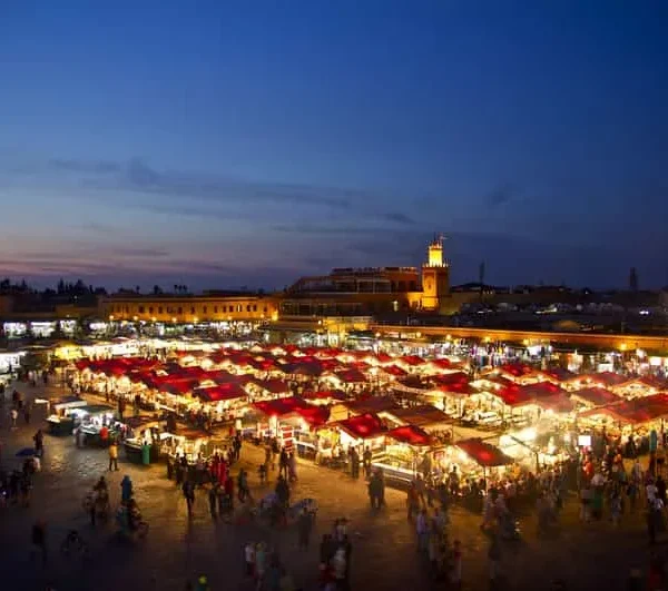 A vibrant night market in Marrakesh, illuminated by colorful lights and bustling with people and stalls.
