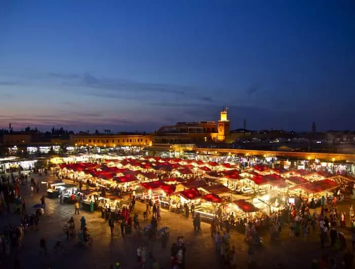 A vibrant night market in Marrakesh, illuminated by colorful lights and bustling with people and stalls.
