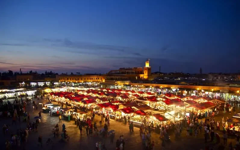A vibrant night market in Marrakesh, illuminated by colorful lights and bustling with people and stalls.