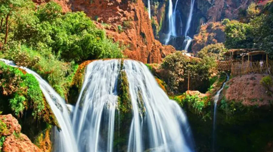 A serene desert waterfall flows beside a quaint hut, surrounded by arid landscape and vibrant natural beauty.