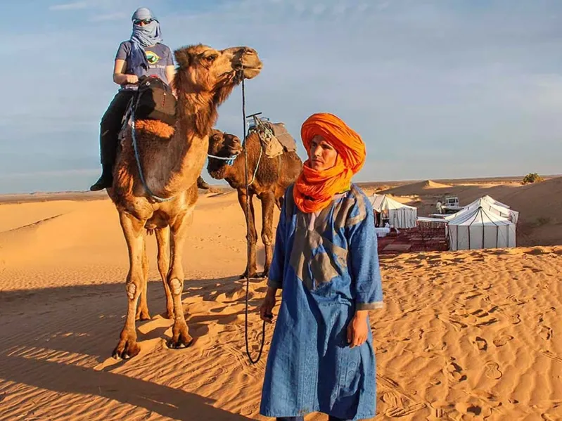 A man in an orange turban and blue dress stands beside two camels, showcasing a scene from a Morocco camel tour.