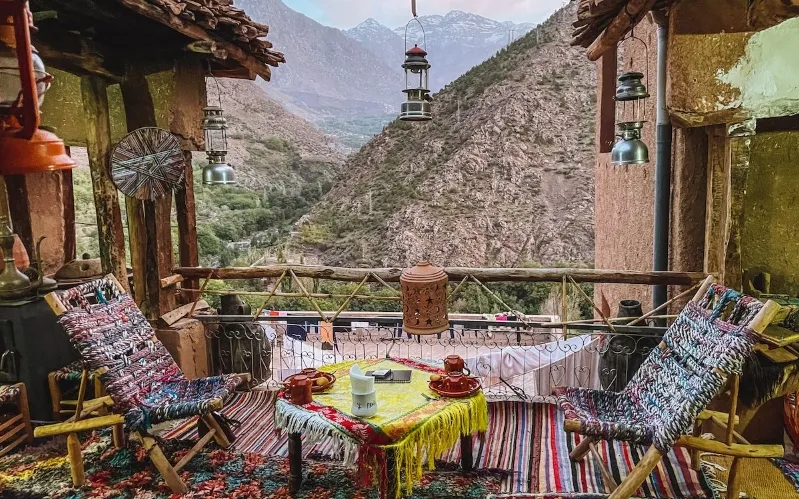 A rustic balcony with colorful woven chairs, a small table with tea settings, hanging lanterns, and a view of mountains and a valley in the background.