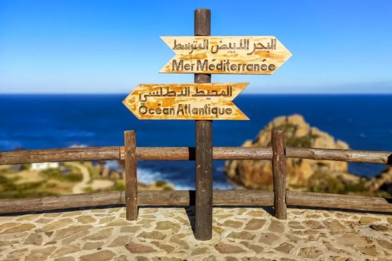 A sign on a pole displaying the text "البحر الأبيض المتوسط Mer Méditerranée المحيط الأطلسى. Ocean Atlantique" with a body of water in the background.