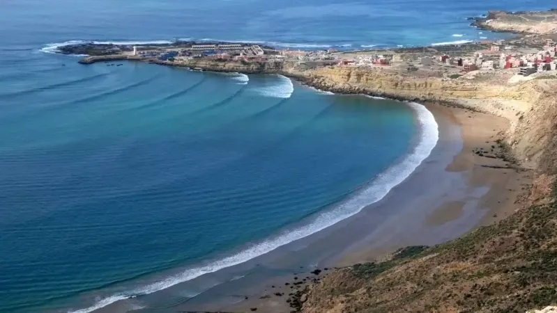 A scenic beach in Morocco featuring a few houses and a cliff, ideal for surf travel enthusiasts. 