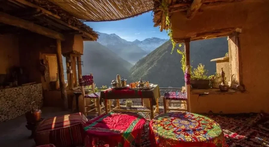 A porch featuring several tables and chairs, surrounded by a scenic mountain landscape. The area includes indoor elements like a kitchen and dining room table, with walls, a ceiling, and flooring visible in the setting.