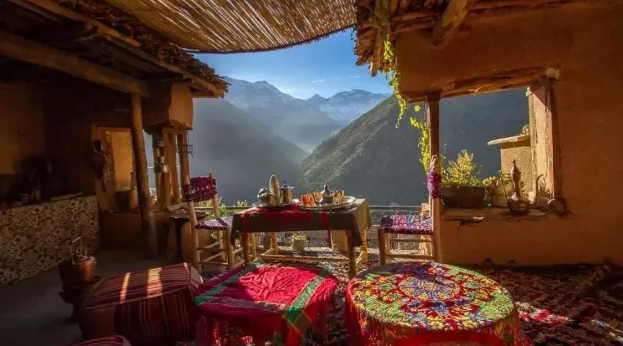 A porch featuring several tables and chairs, surrounded by a scenic mountain landscape. The area includes indoor elements like a kitchen and dining room table, with walls, a ceiling, and flooring visible in the setting.