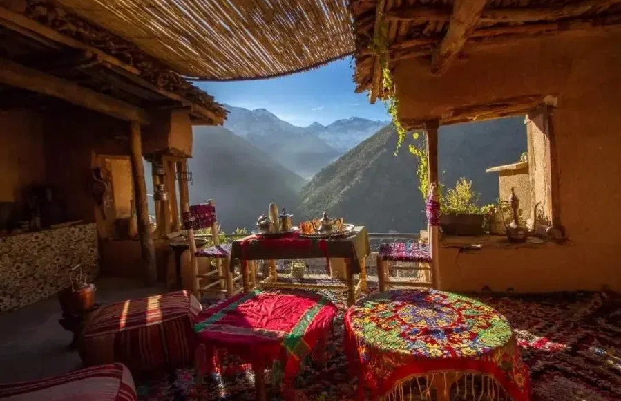 A porch featuring several tables and chairs, surrounded by a scenic mountain landscape. The area includes indoor elements like a kitchen and dining room table, with walls, a ceiling, and flooring visible in the setting.