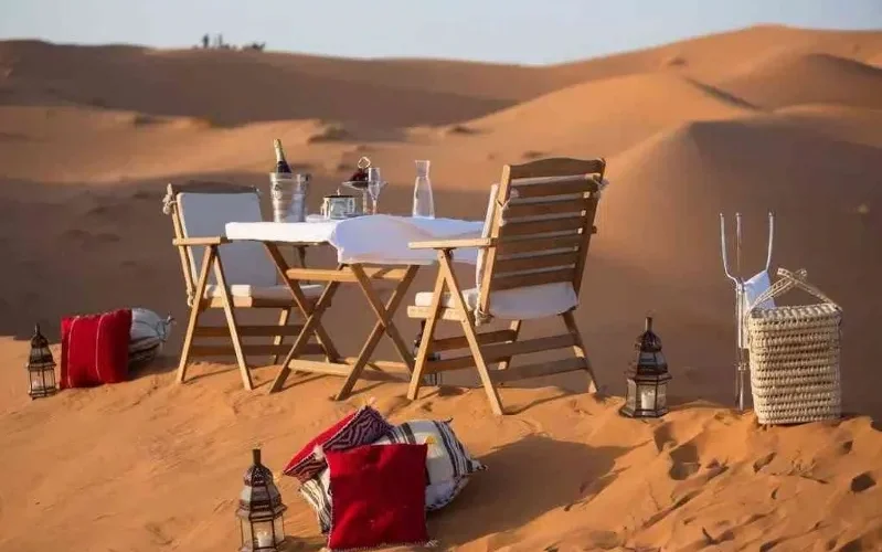 A beautifully set dinner table in the Moroccan desert, surrounded by sand dunes under a starry sky.