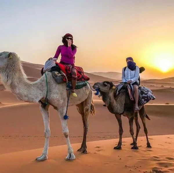 Two people riding camels through the Moroccan desert at sunset, showcasing a vibrant sky and sandy dunes.
