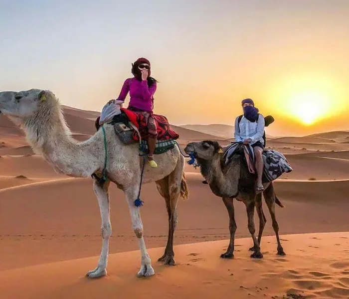 Two people riding camels through the Moroccan desert at sunset, showcasing a vibrant sky and sandy dunes.