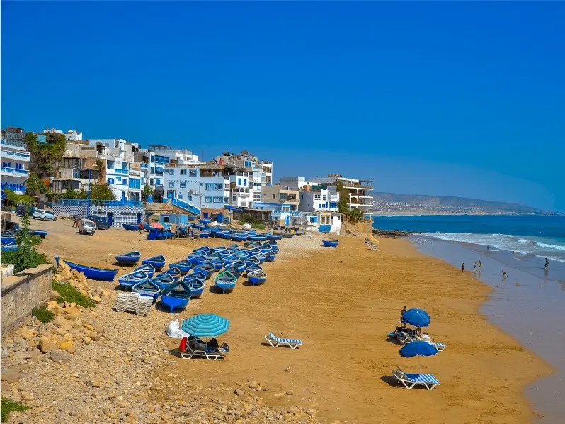 A beach in Morocco featuring blue umbrellas and chairs, ideal for surf travel and relaxation by the ocean. 