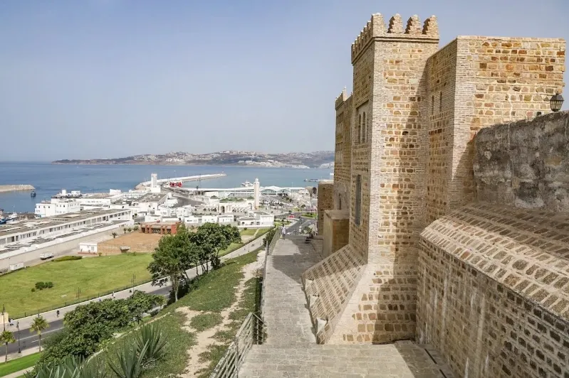 A stone building stands prominently with a body of water visible in the background under a clear sky.While Day Trip to Morocco From Spain