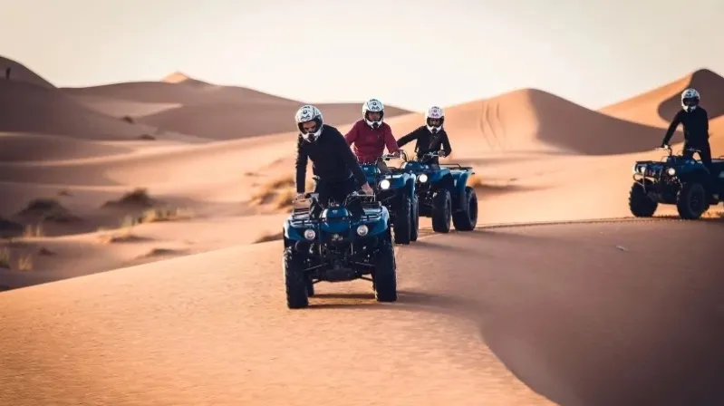 Four people riding ATVs through the Moroccan desert, showcasing the vast sandy landscape and clear blue sky. 