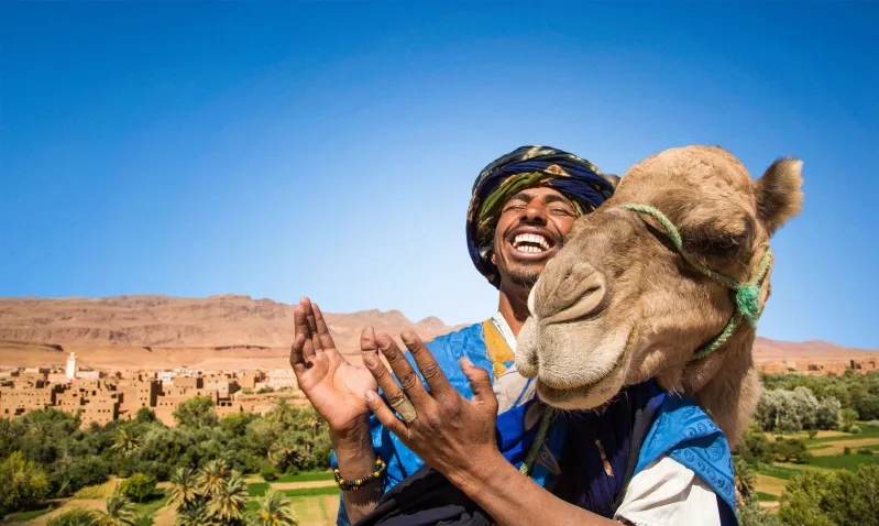 A man in a turban and blue dress holds a camel, promoting Morocco trip packages.