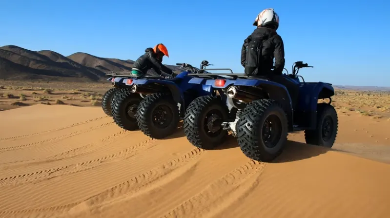 Two people riding four wheelers across a sandy desert landscape, promoting Morocco tour packages.