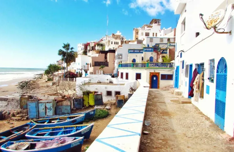 A scenic beach in Agadir featuring blue and white buildings alongside colorful boats.visit Agadir