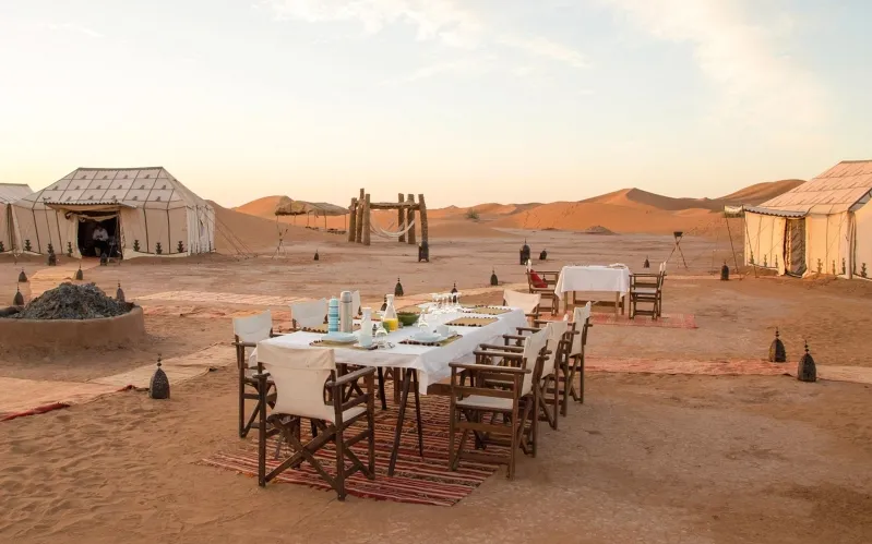 A table set in the Moroccan desert, surrounded by tents and chairs, creating a cozy outdoor dining atmosphere.