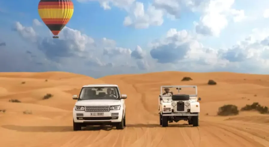 Two vehicles drive through a desert landscape with a colorful hot air balloon floating in the background, promoting Morocco custom tours.