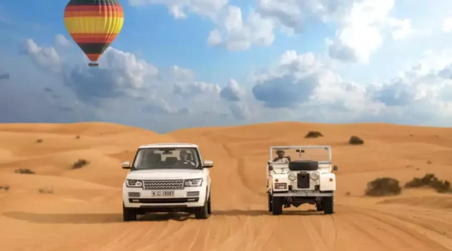 Two vehicles drive through a desert landscape with a colorful hot air balloon floating in the background, promoting Morocco custom tours.