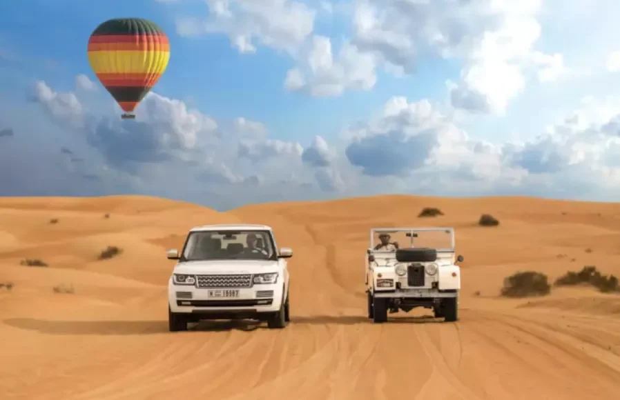 Two vehicles drive through a desert landscape with a colorful hot air balloon floating in the background, promoting Morocco custom tours.