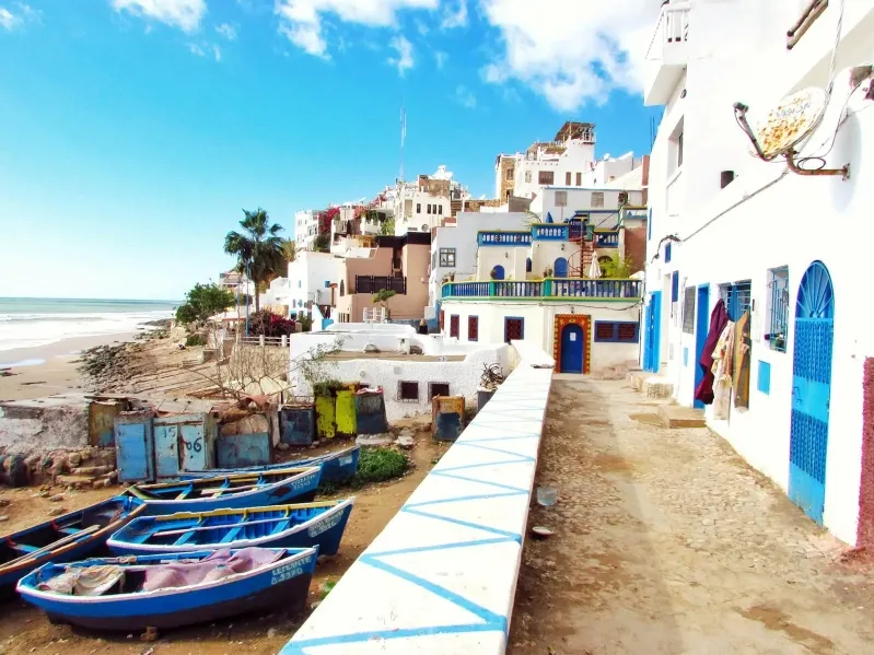 A row of colorful boats on the beach beside a white building, showcasing a scenic view in Morocco.
