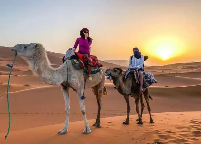 Two people riding camels in the desert during sunset, part of the Fes to Marrakech Desert Tour.