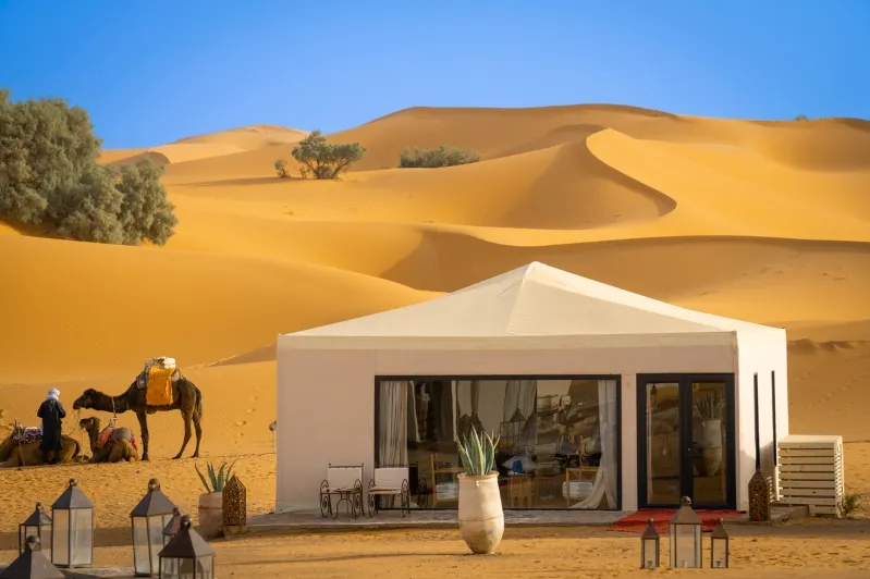 A desert scene featuring a tent, camels, and people, highlighting the Marrakech to Fes Desert Tour experience. 