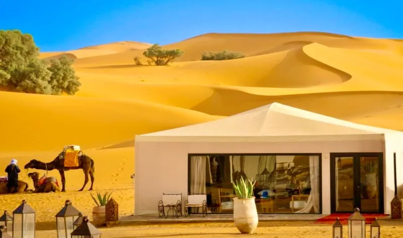 A luxury tent in the desert with a camel and a man, showcasing private tours in Morocco's stunning landscape.