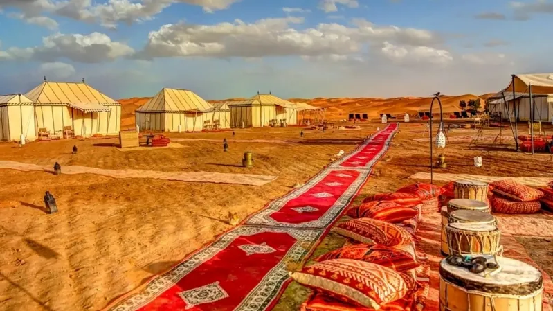 A serene view of a Moroccan desert luxury camp with tents and a decorative carpet set against the arid landscape.