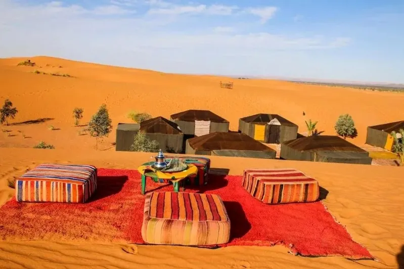 A vibrant rug and a table set in a luxurious desert camp in Merzouga, Morocco, surrounded by sandy dunes.
